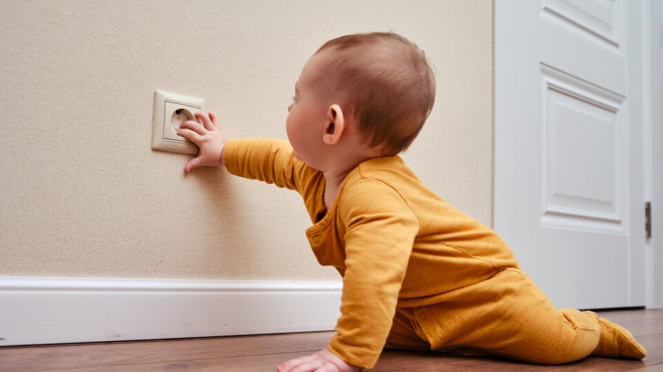 Baby toddler reaches into the electrical outlet on the home wall with his hand. Danger and protection of child fingers from electric shock, aged 6-11 months
